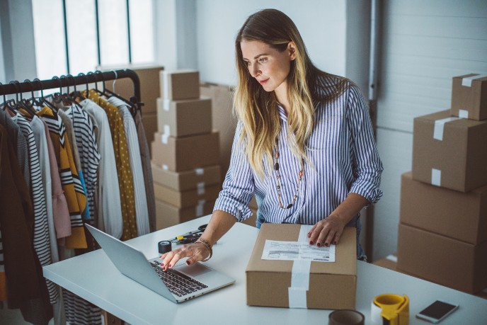 Operating a business from home - woman with laptop and parcel beside clothes rail