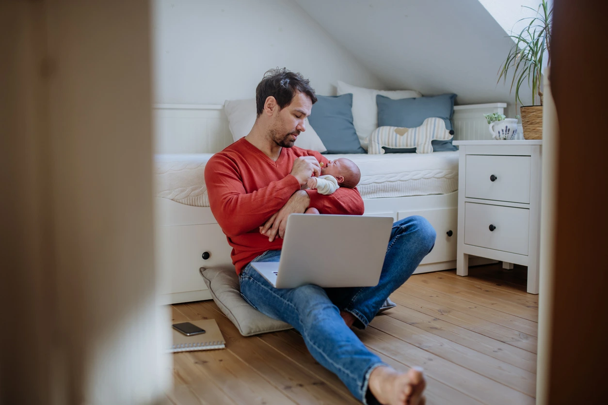 A father sitting on the floor holding a baby with a laptop on his lap