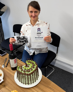 a picture of a woman with dark hair holding a certificate behind a prize winning green and brown cake